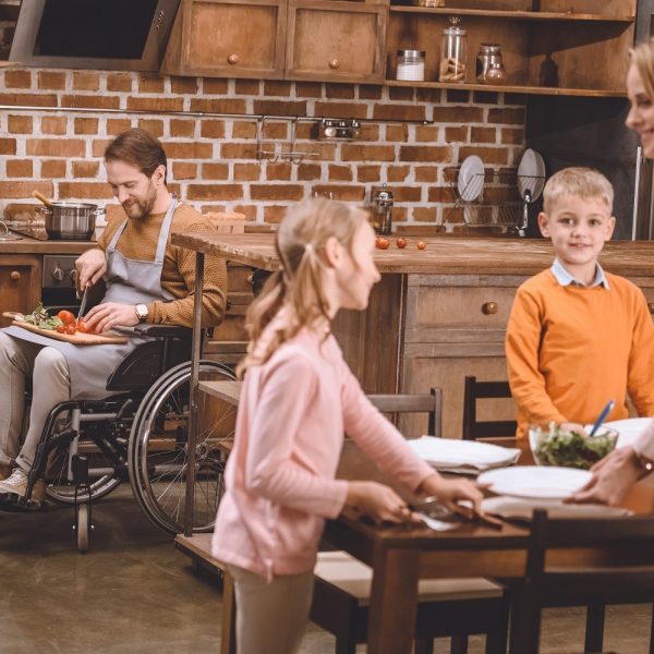 A woman and two children set the table while a man, seated in wheelchair, chops some vegetables