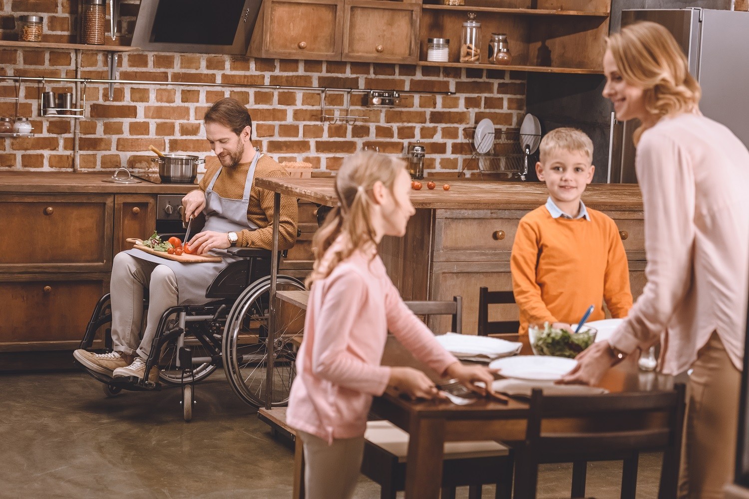 A woman and two children set the table while a man, seated in wheelchair, chops some vegetables