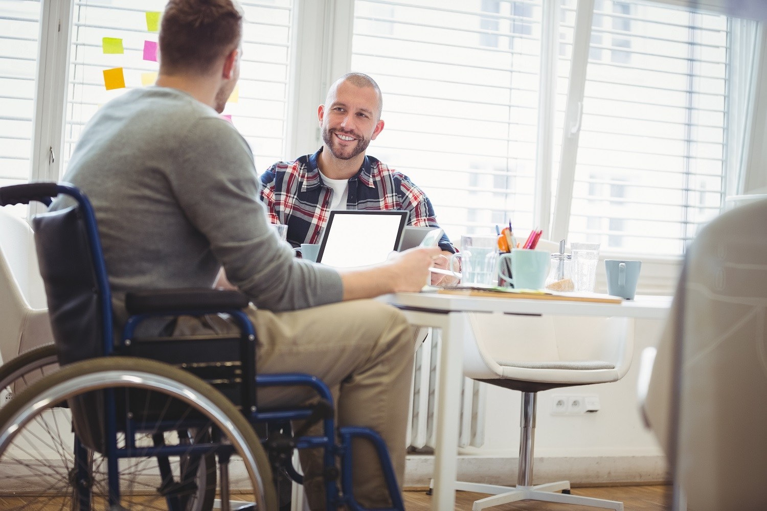 A man in a wheelchair in an office setting in conversation with another man at a desk