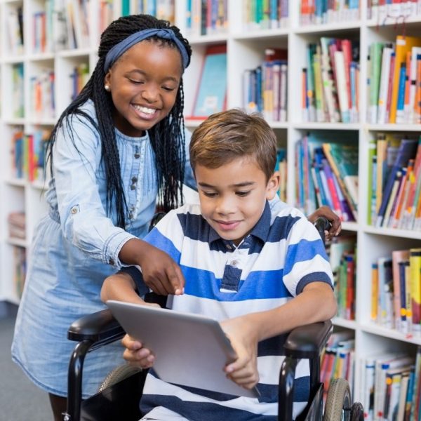 Two children in a library. One is holding an iPad and sitting in a wheelchair