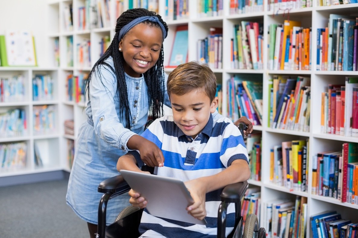 Two children in a library. One is holding an iPad and sitting in a wheelchair