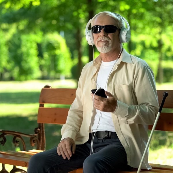 man wearing dark glasses, using headphones with his phone, seated at a park bench