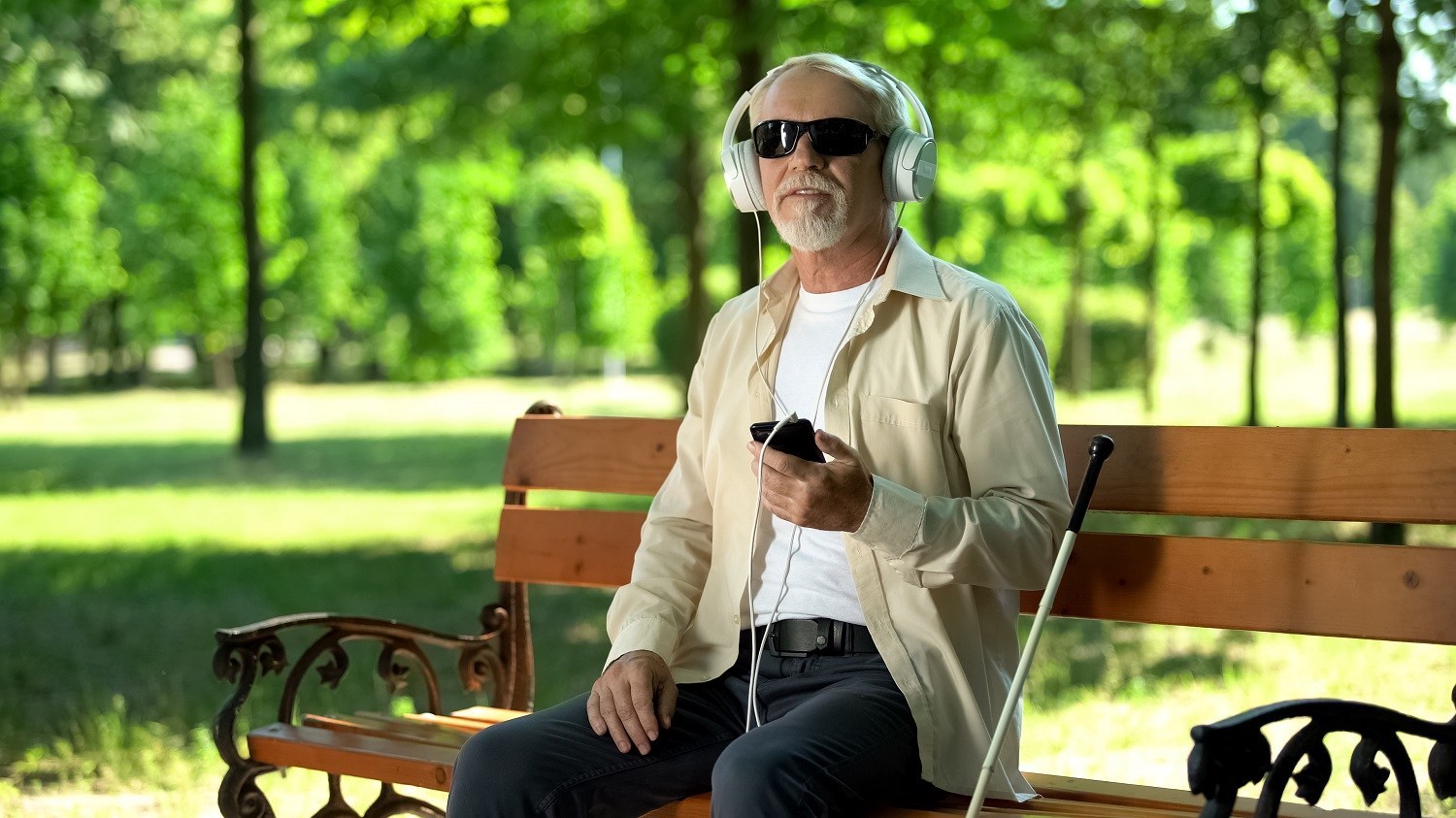 man wearing dark glasses, using headphones with his phone, seated at a park bench