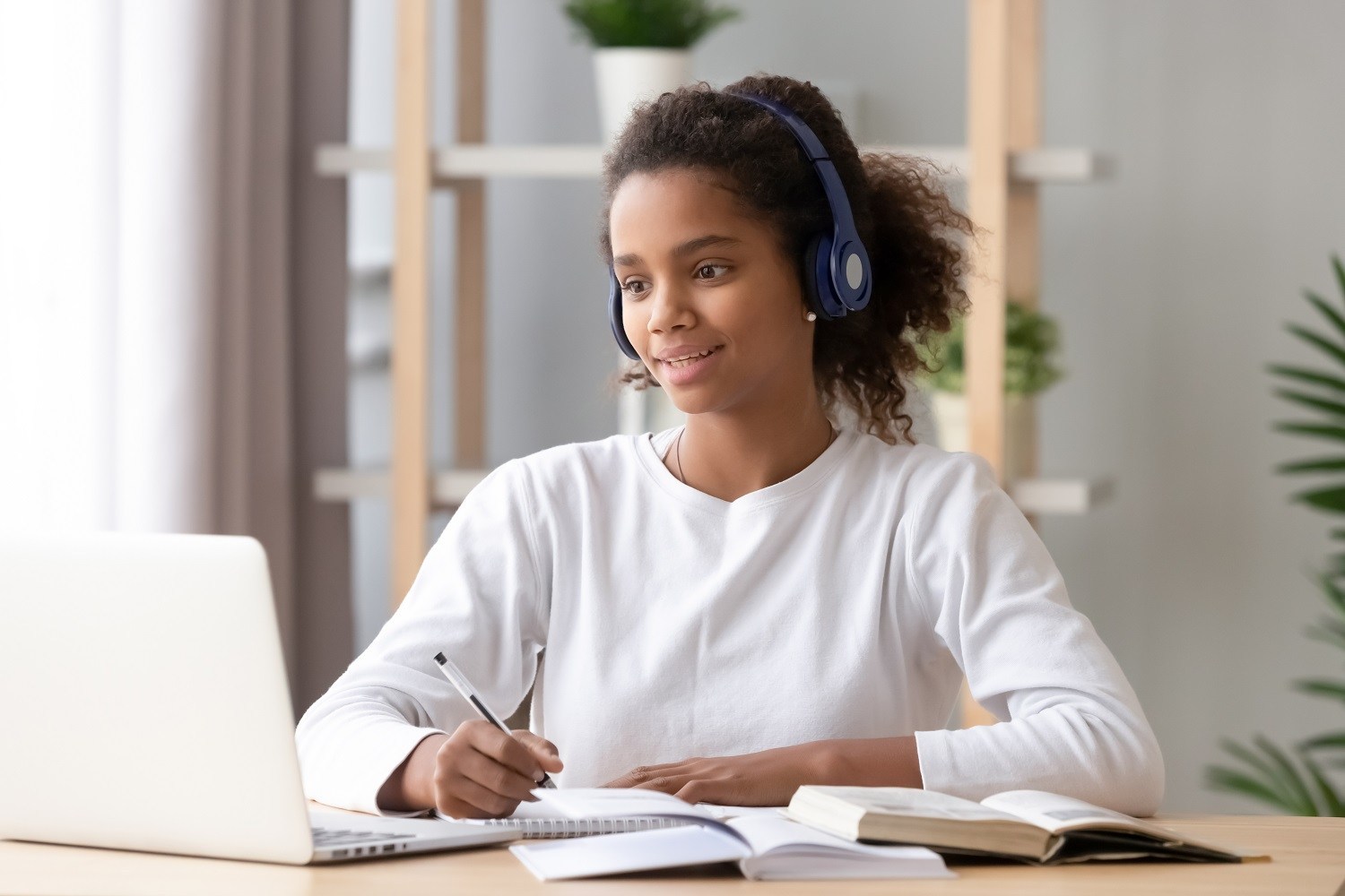 student using headphones and looking at laptop, taking notes in notebook