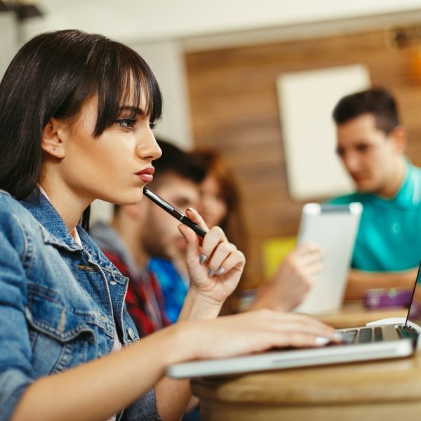 A classroom setting. In the foreground, a student is using a laptop