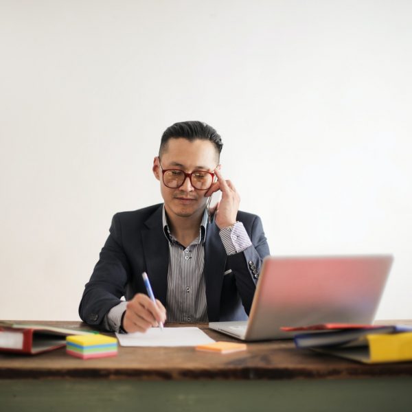 man seated at a desk, speaking on the phone and writing notes by hand