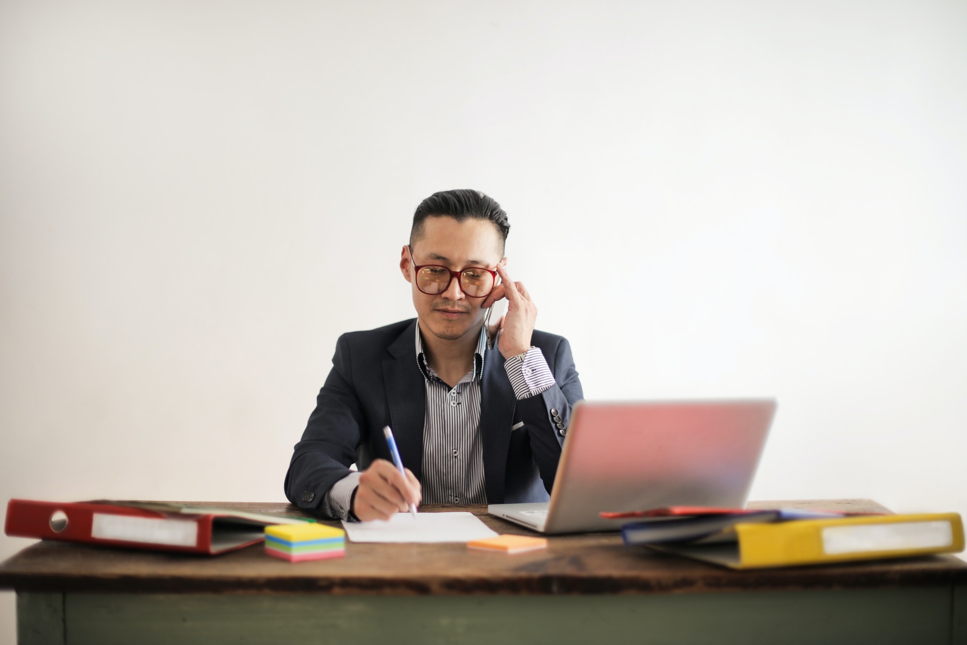 man seated at a desk, speaking on the phone and writing notes by hand