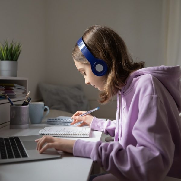 a student using headphones while working on her laptop and writing in a notebook