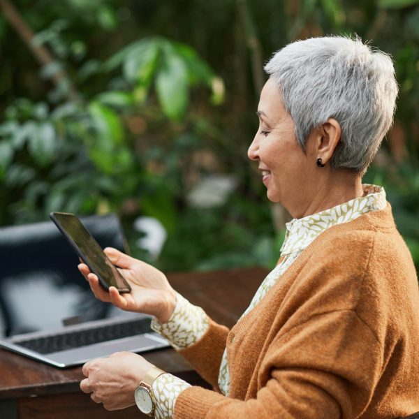 A woman seated outdoors, using a phone. Her laptop is on a table