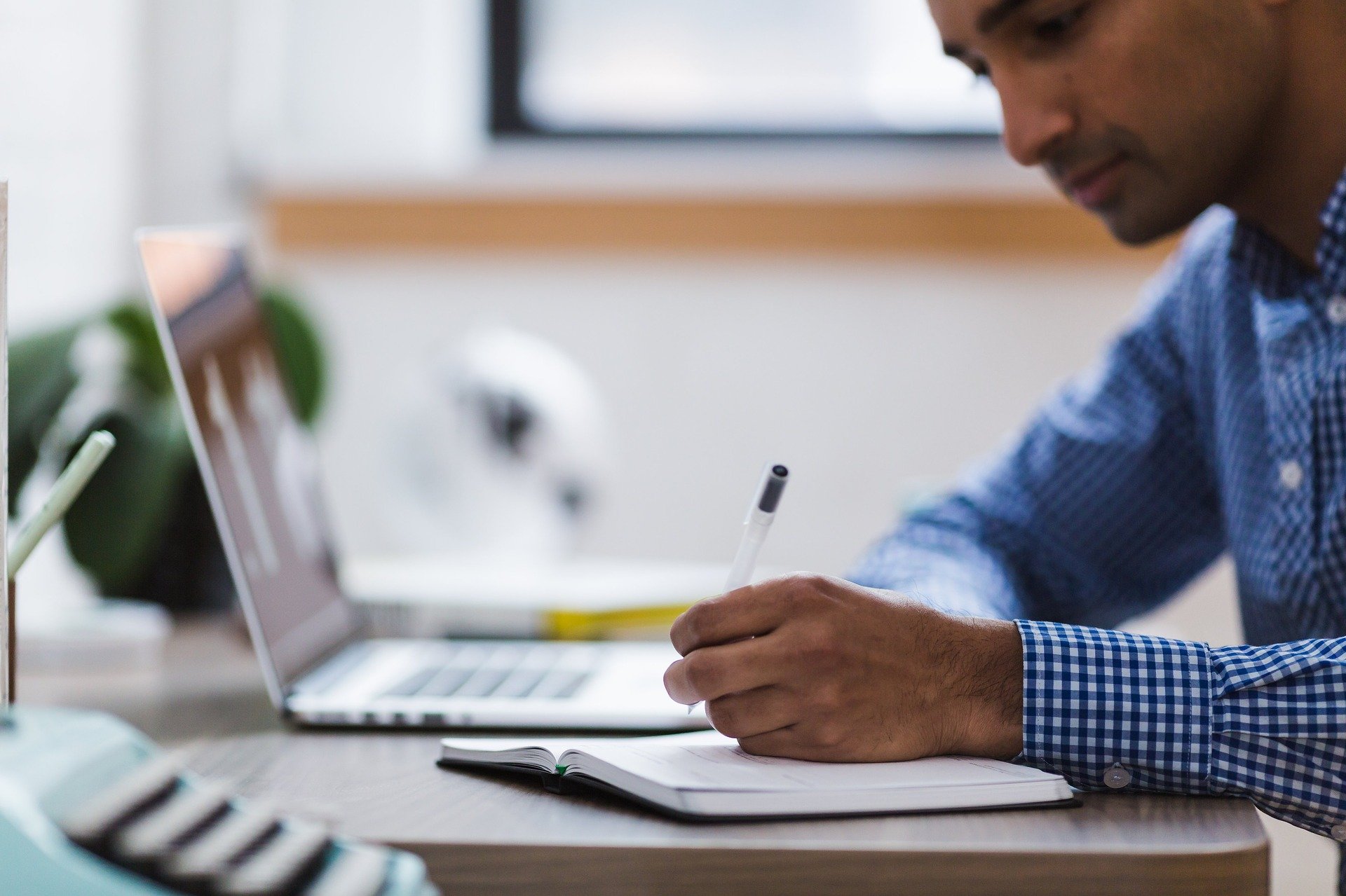 man working at a laptop, writing notes in a book