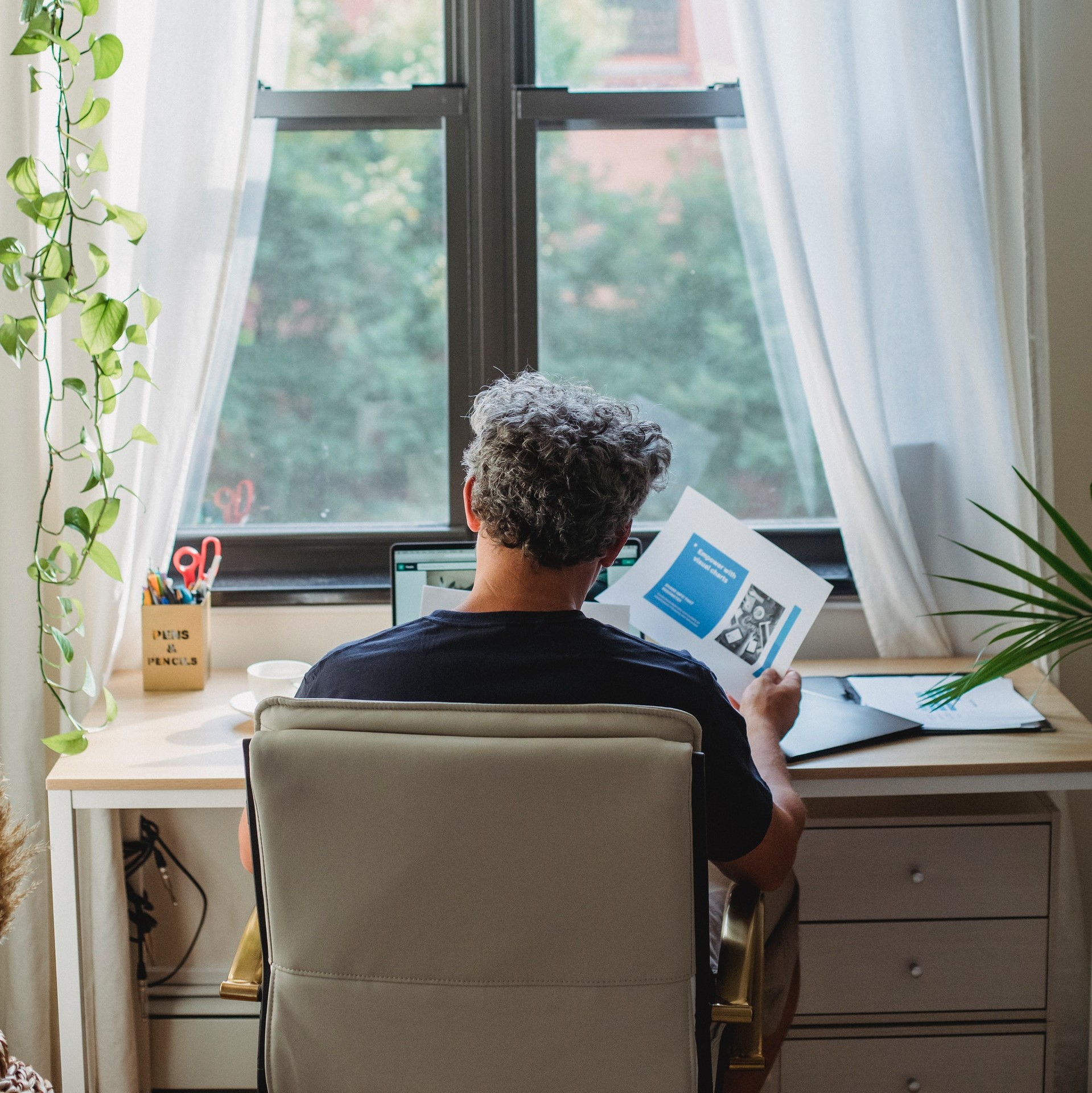 stock image showing back view of a man seated at a desk, browsing some papers with a laptop on the desk