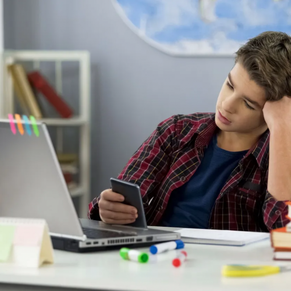 A student seated at a desk, using a smartphone. In the foreground is a laptop and a stack of books