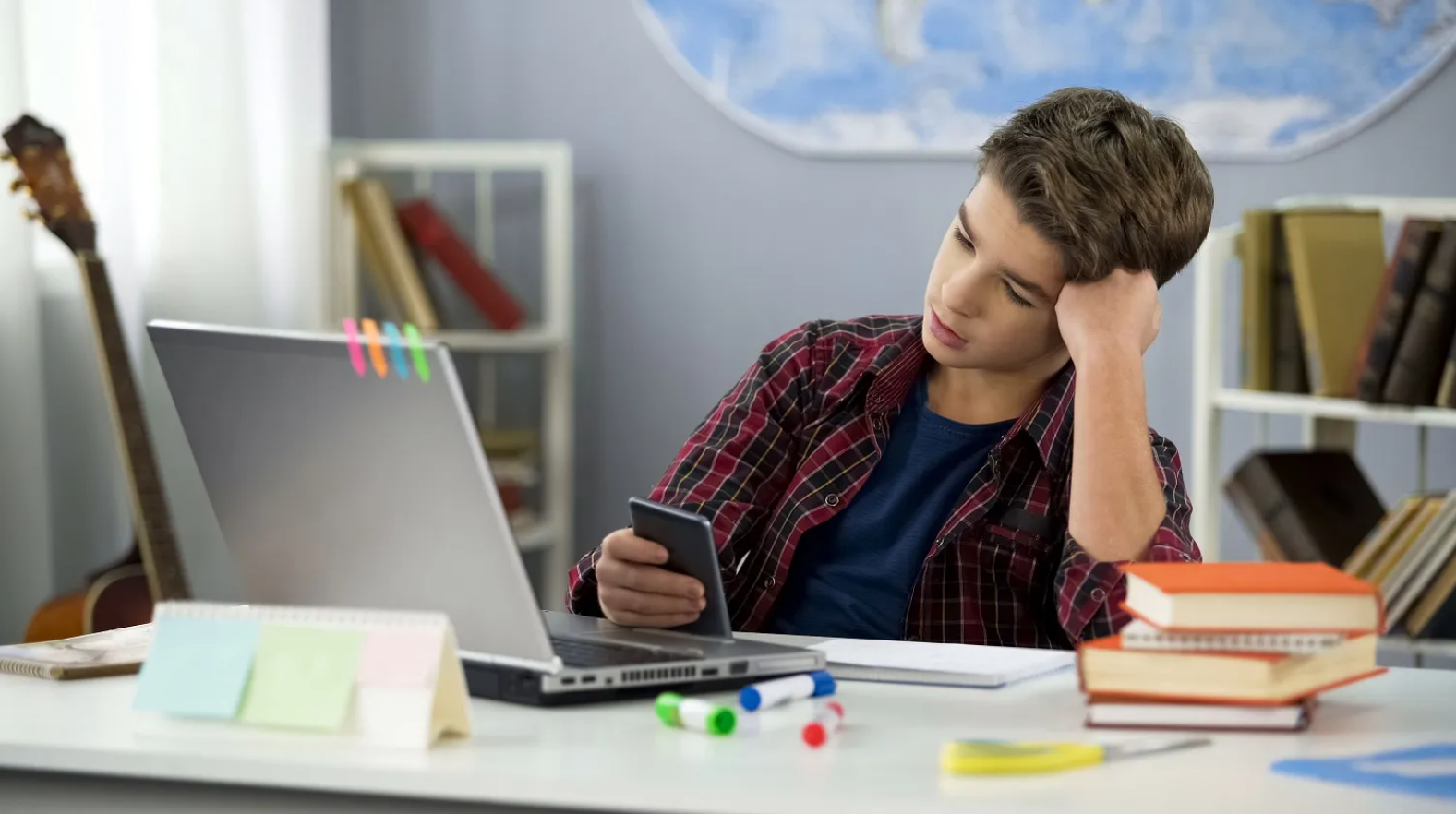 A student seated at a desk, using a smartphone. In the foreground is a laptop and a stack of books