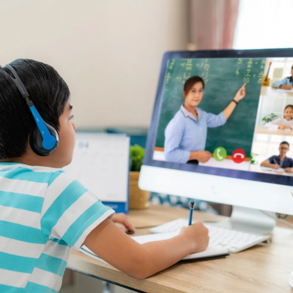 A child wearing headphones, seated at a desk, watching a teacher gesturing towards a blackboard