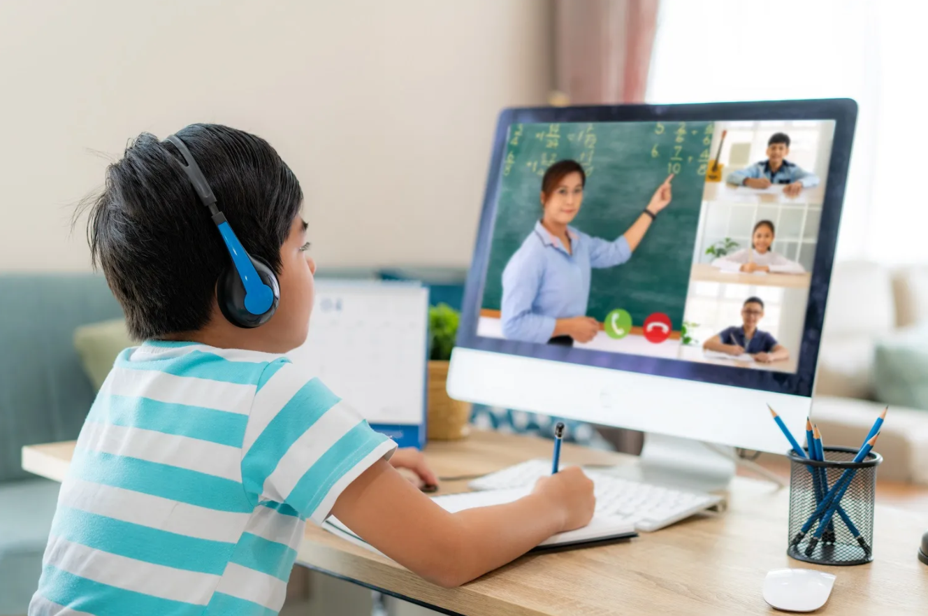 A child wearing headphones, seated at a desk, watching a teacher gesturing towards a blackboard