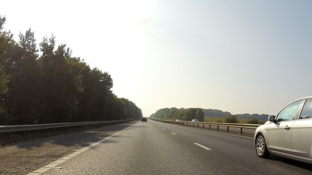 A lone car driving along a tree-lined road under a clear sky