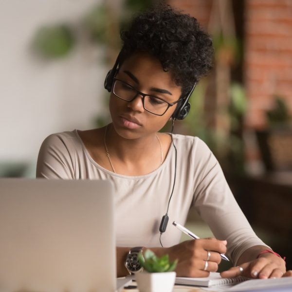 a woman wearing headphones writing down notes with a pen while looking at a laptop