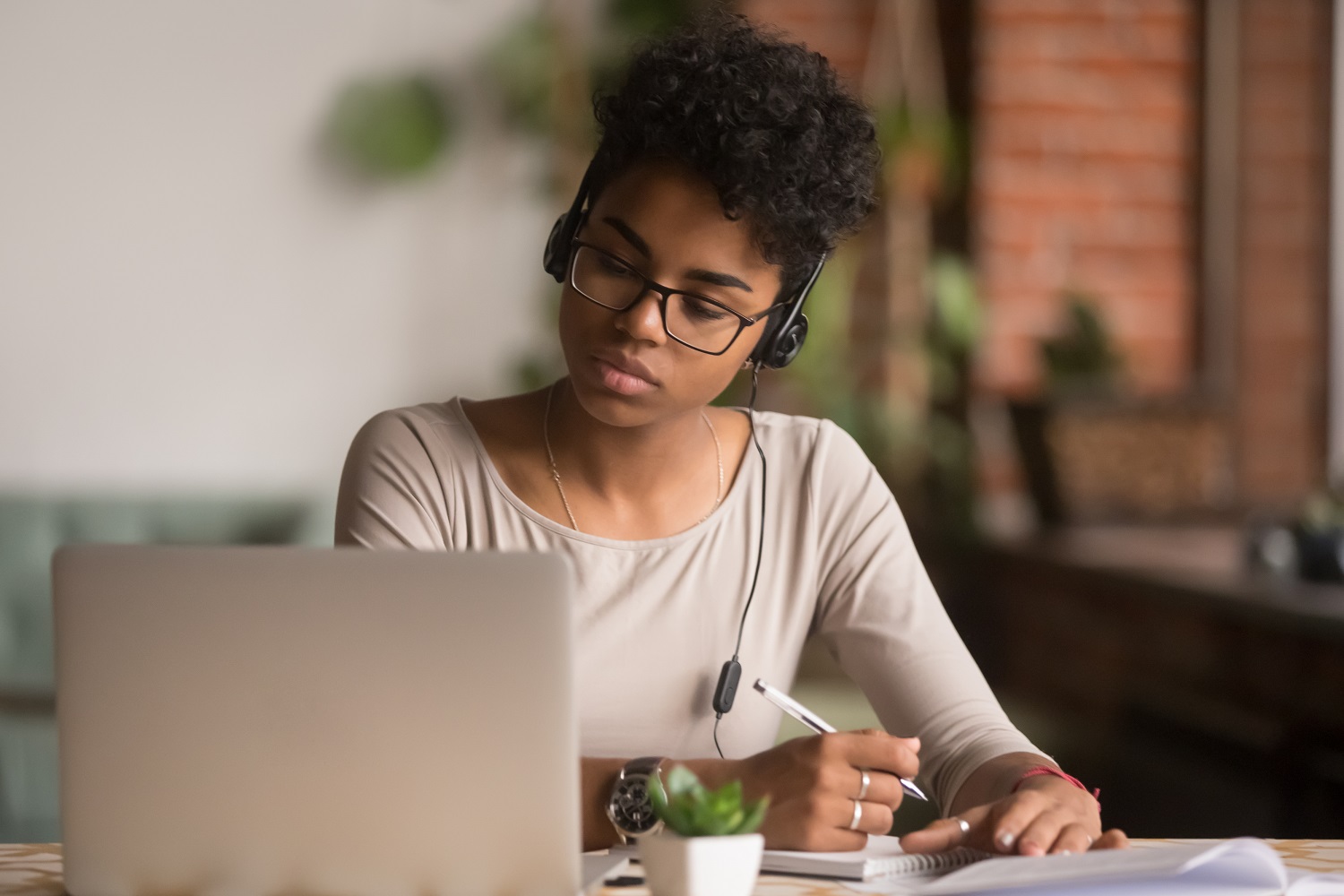 a woman wearing headphones writing down notes with a pen while looking at a laptop