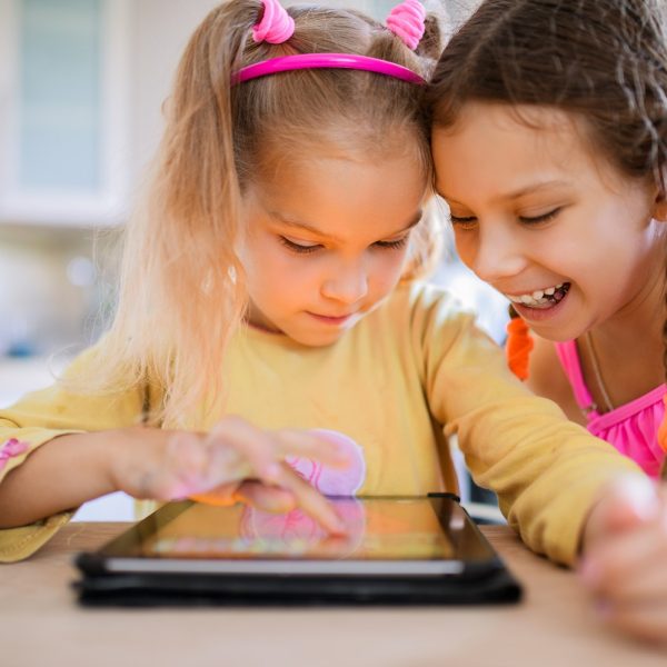 two children are using a tablet, one is scrolling, while the other is watching with a smile