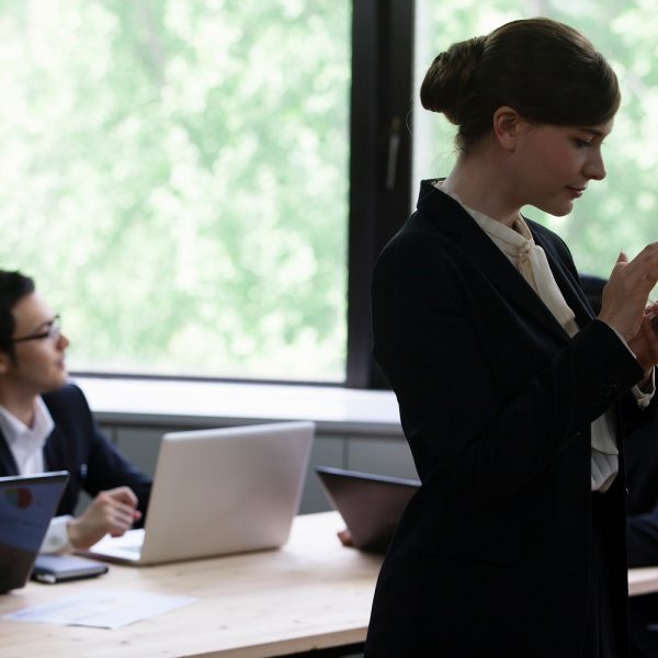 a man sits at a desk working on a laptop in the background, while a woman, who is standing, scrolls her phone