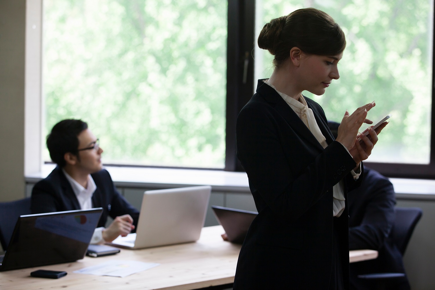 a man sits at a desk working on a laptop in the background, while a woman, who is standing, scrolls her phone