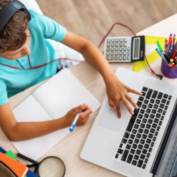 a young boy studying at home with a laptop and notebook, with various school supplies on the desk