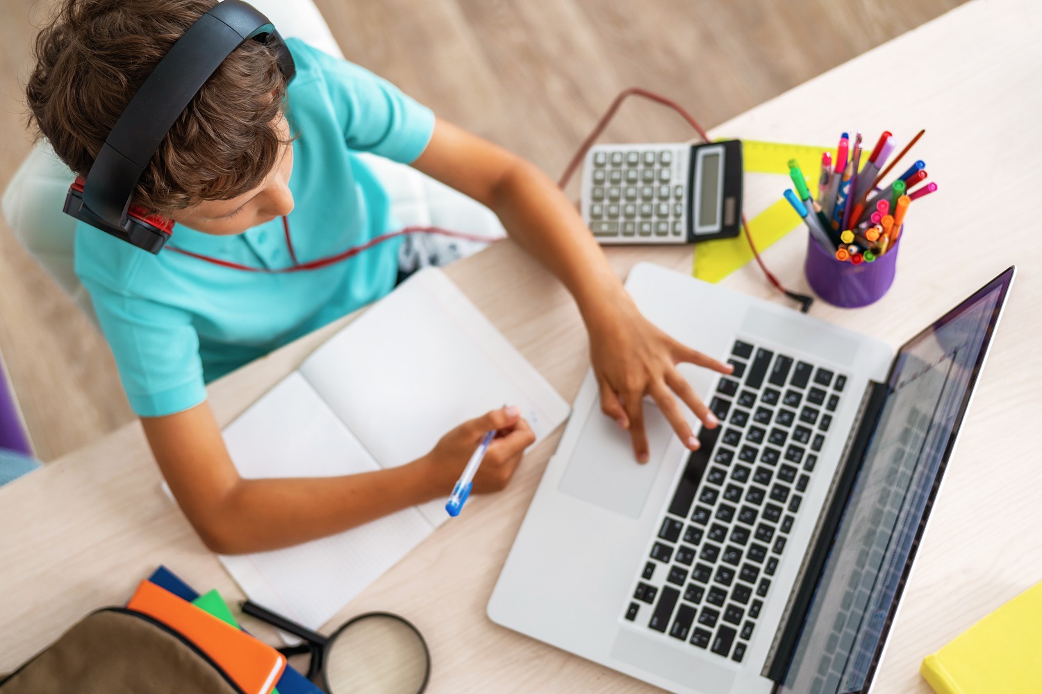 a young boy studying at home with a laptop and notebook, with various school supplies on the desk