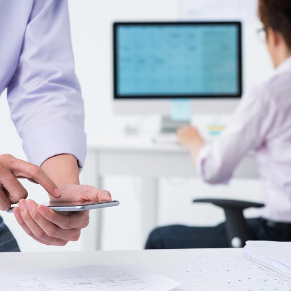 a person looking at a phone while standing over a table, a person is on a computer in the background