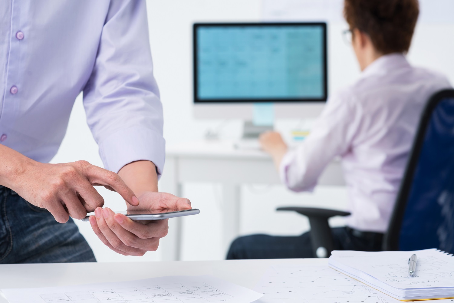 a person looking at a phone while standing over a table, a person is on a computer in the background