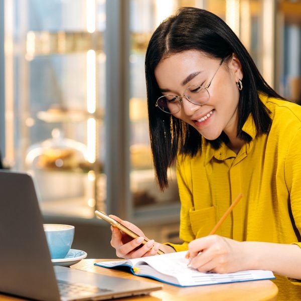 a woman in a cafe holding her phone in one hand and writing in her notebook in the other, with her laptop open