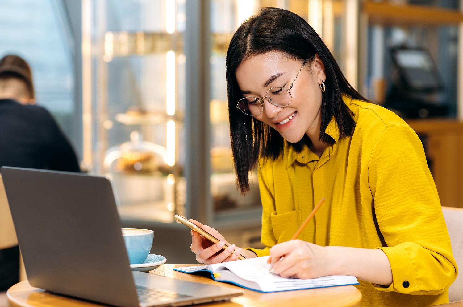 a woman in a cafe holding her phone in one hand and writing in her notebook in the other, with her laptop open