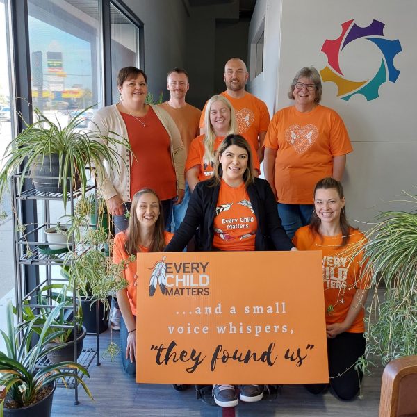 Neil Squire Regina staff wearing orange shirts and a sign that says, "Every Child Matters. ...A small voice whispers, "They found us"