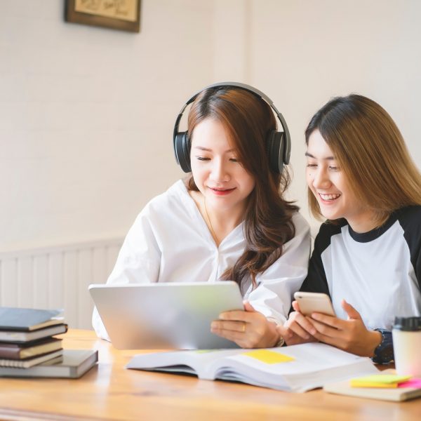 two women working together, one holding a tablet and one holding a phone, with books on the table