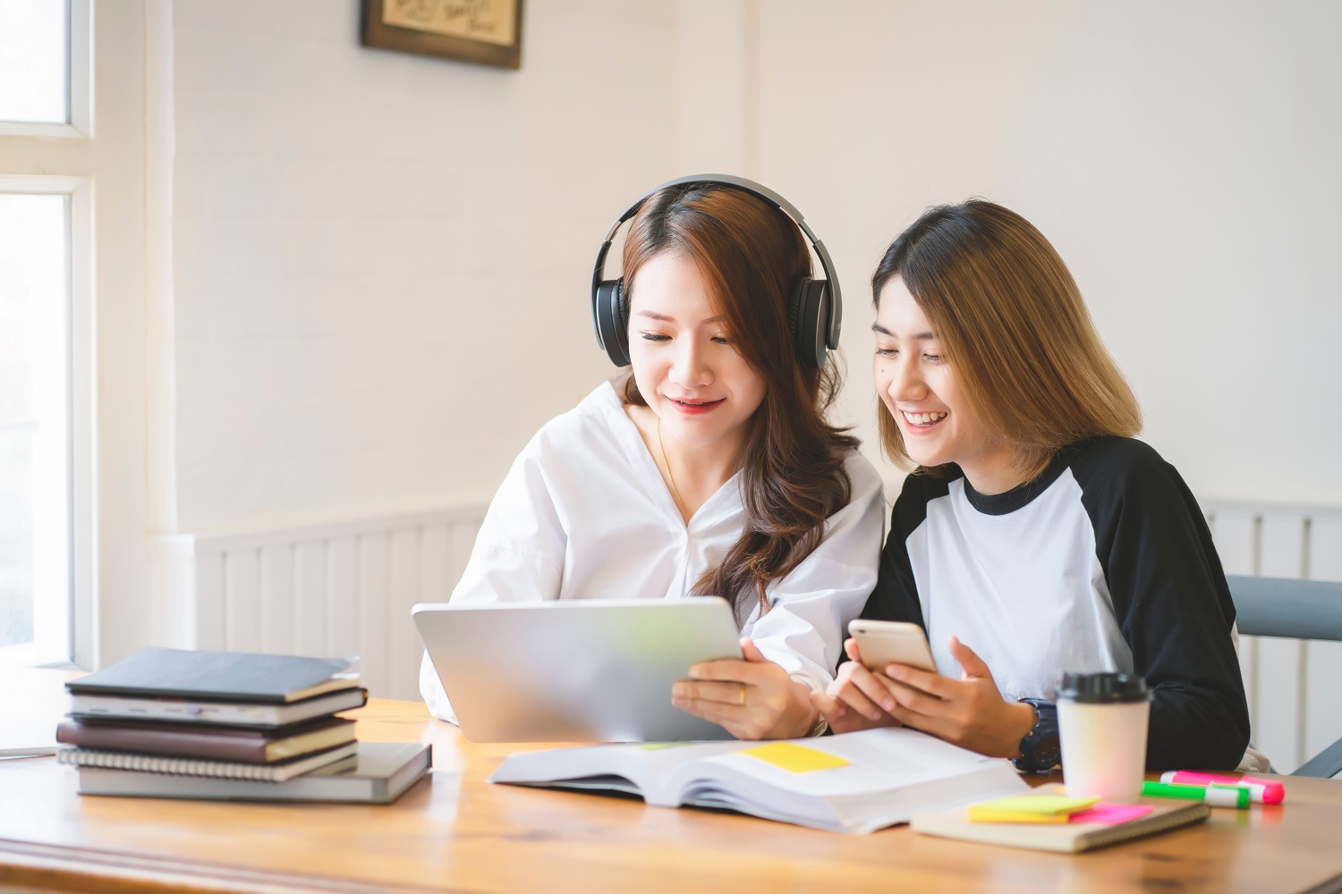 two women working together, one holding a tablet and one holding a phone, with books on the table