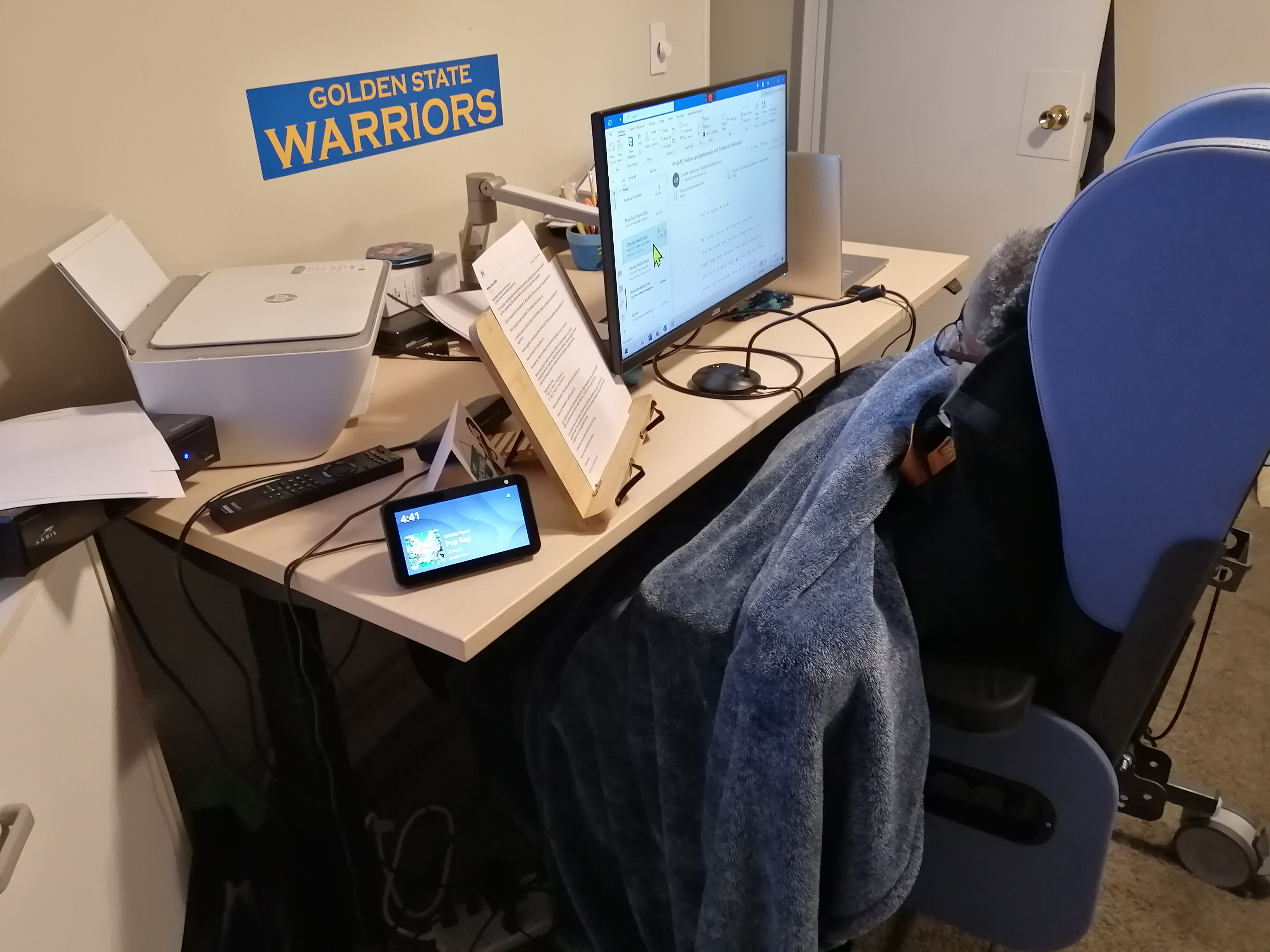 Andrea at her desk in a wheelchair. Her desk is a sit-stand desk and she has a microphone to speak into the computer.