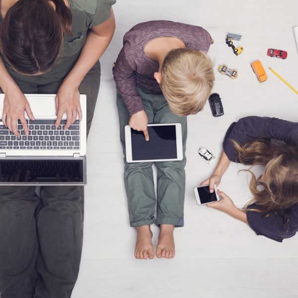 a mom using a laptop sitting next to a young boy using a tablet and a young girl using a smartphone