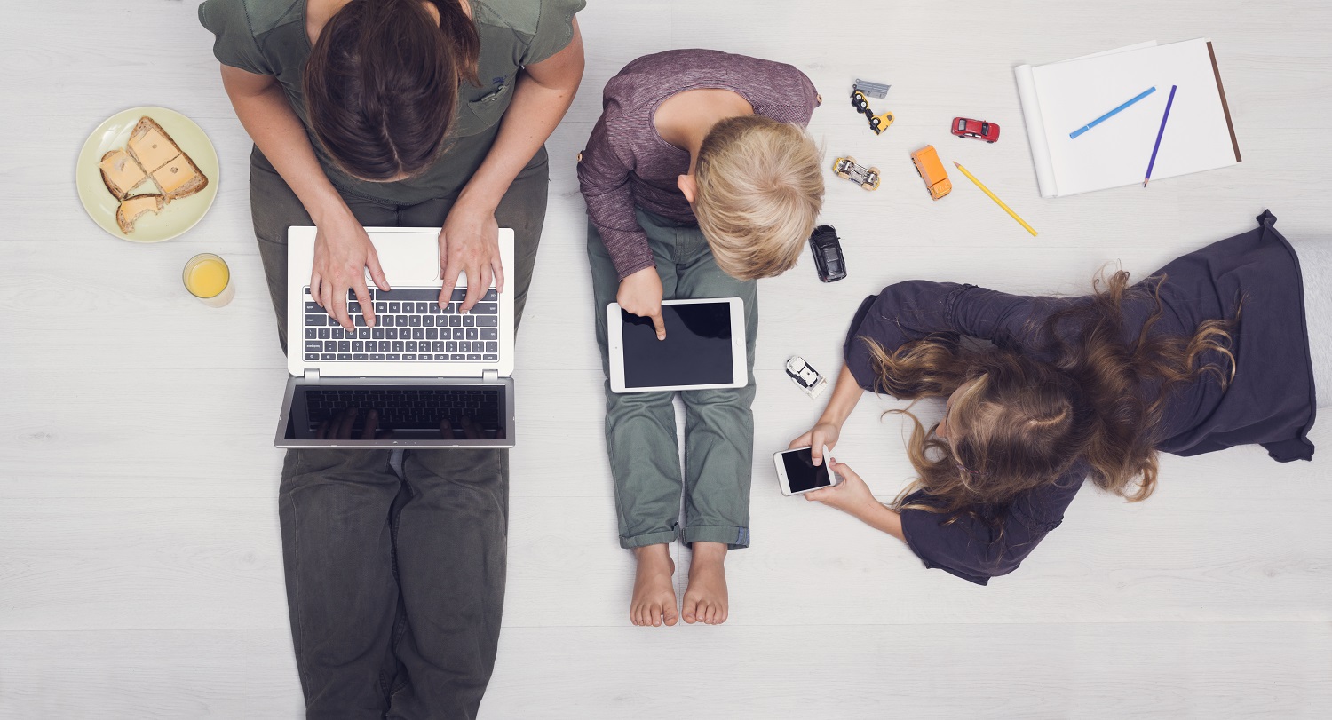 a mom using a laptop sitting next to a young boy using a tablet and a young girl using a smartphone