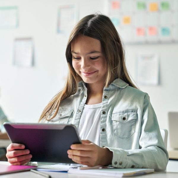 A young girl in a classroom smiles while using a tablet.