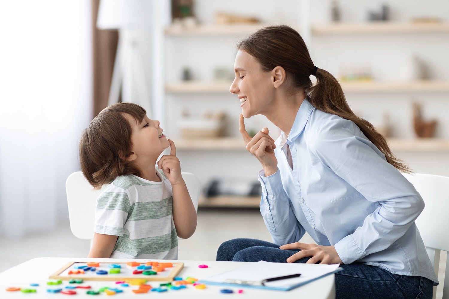 An adult women and a little girl smile at each other while each touching their chins in a speech exercise.