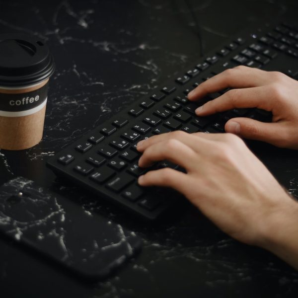 A person types on a keyboard with a cup of coffee next to it.