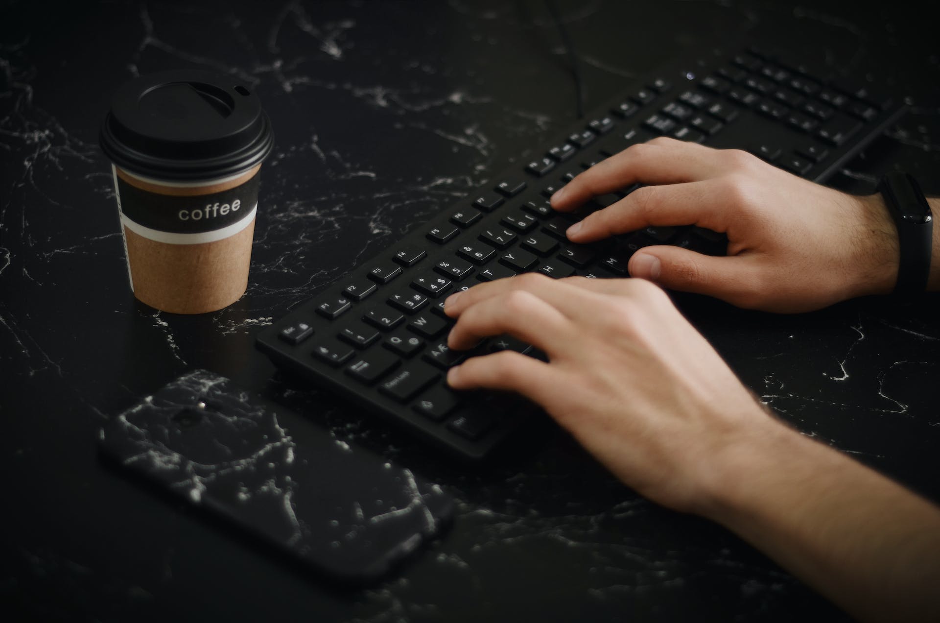 A person types on a keyboard with a cup of coffee next to it.