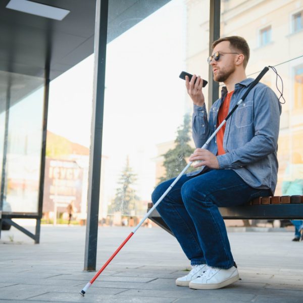 A blind man wearing sunglasses and a white cane speaks into his phone while sitting on a bench.