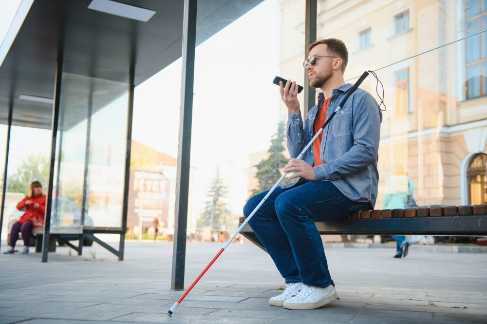 A blind man wearing sunglasses and a white cane speaks into his phone while sitting on a bench.