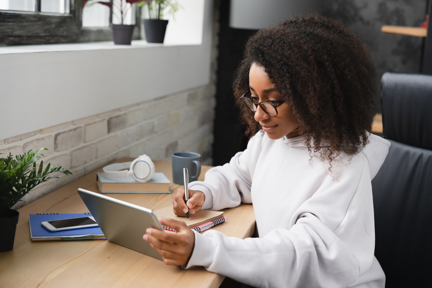 A woman writes on a notepad while watching something on a tablet.