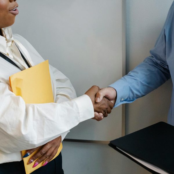 A woman holding a folder shakes hands with a man in a suit holding a book.