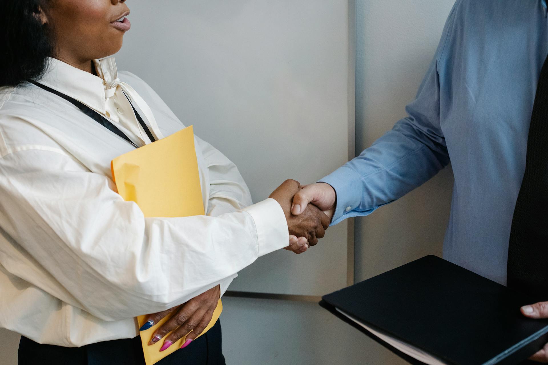 A woman holding a folder shakes hands with a man in a suit holding a book.