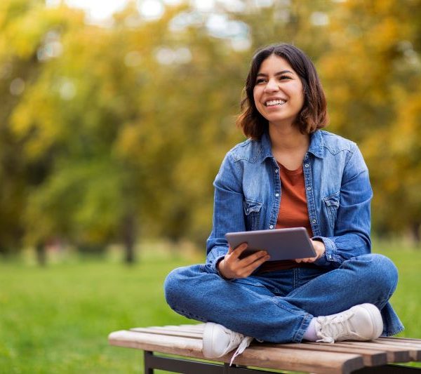 A woman sits cross-legged on a bench outside holding a tablet.