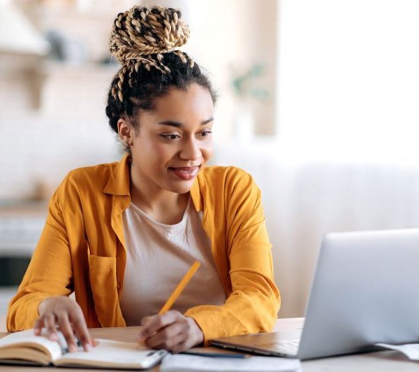 A woman writes into a notebook while using the laptop.