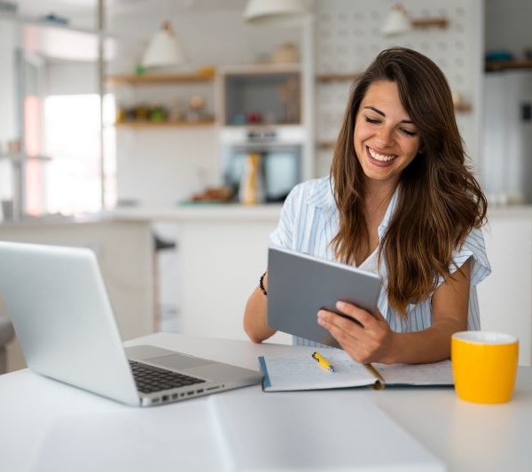 A woman sitting near a notepad pen, uses a tablet while also having a laptop open.