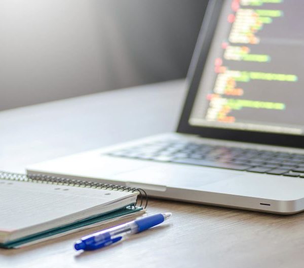 A laptop being used for coding, with a notebook and pen in front of it.